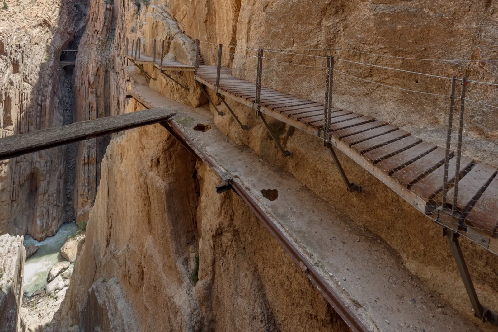 Walker's-eye view along the Caminito del Rey wooden plank boardwalk as it runs ahead and bends around an ochre cliff, with the crumbling original concrete ledge visible just below it