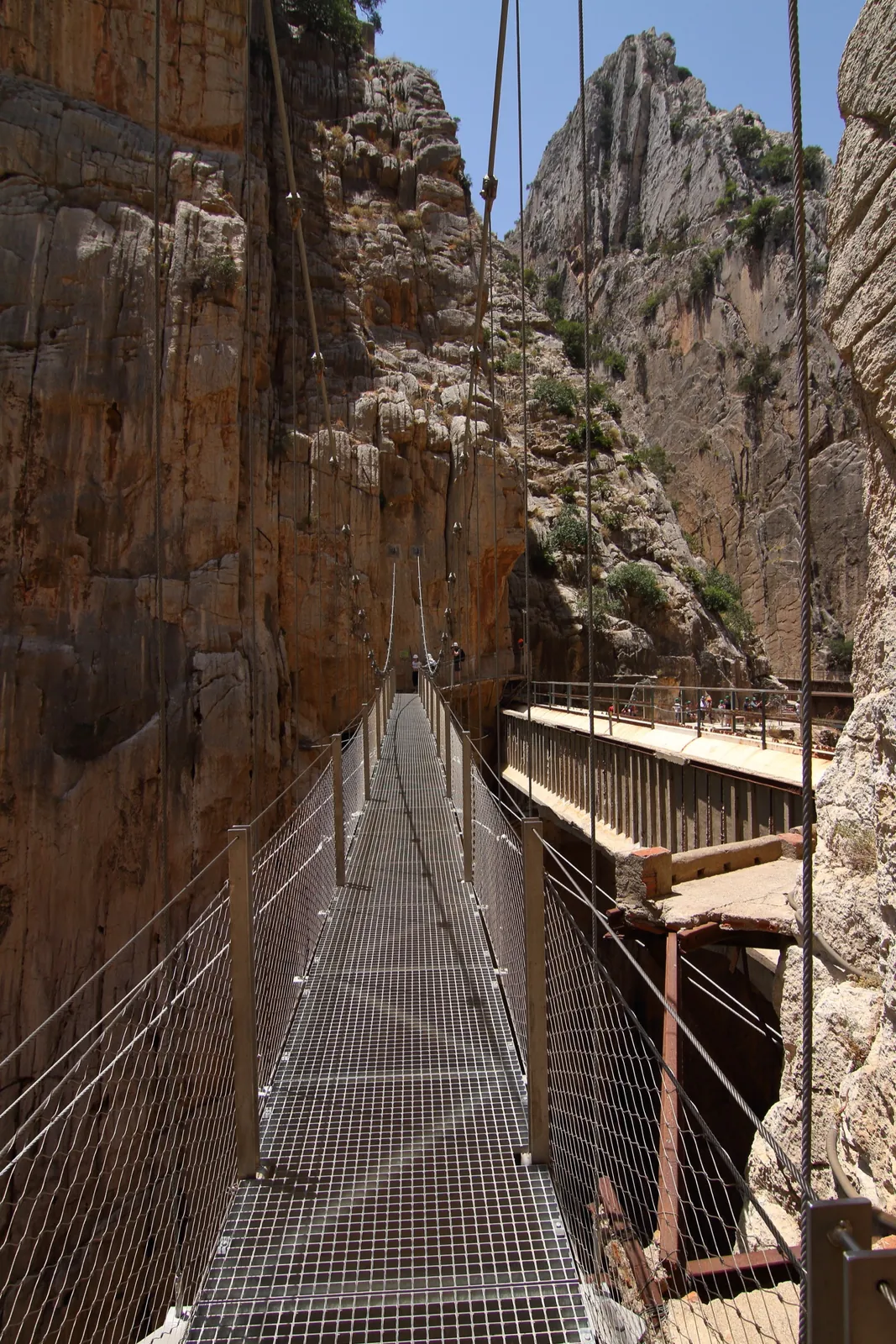 Point-of-view along the Caminito del Rey suspension footbridge, a narrow steel-mesh-deck cable bridge spanning the gorge with sheer cliffs rising on both sides