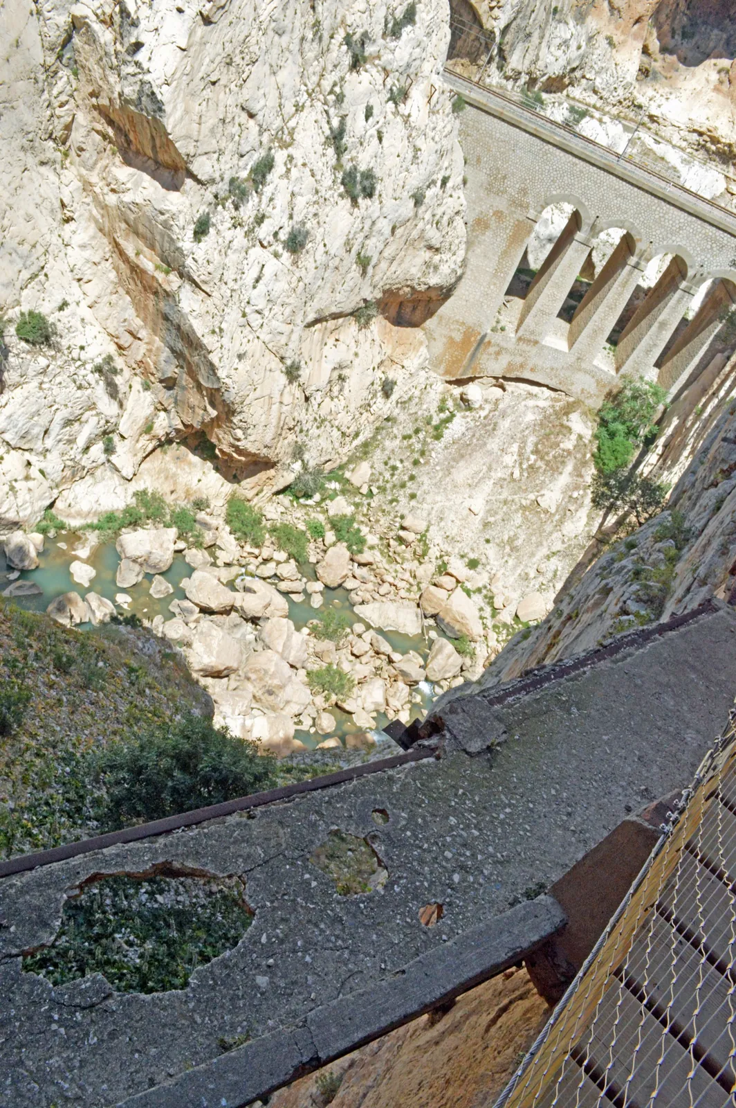 The derelict pre-2015 Caminito del Rey path: a broken concrete cantilever ledge with a collapsed, holed section and no railing, above the river and an old arched aqueduct